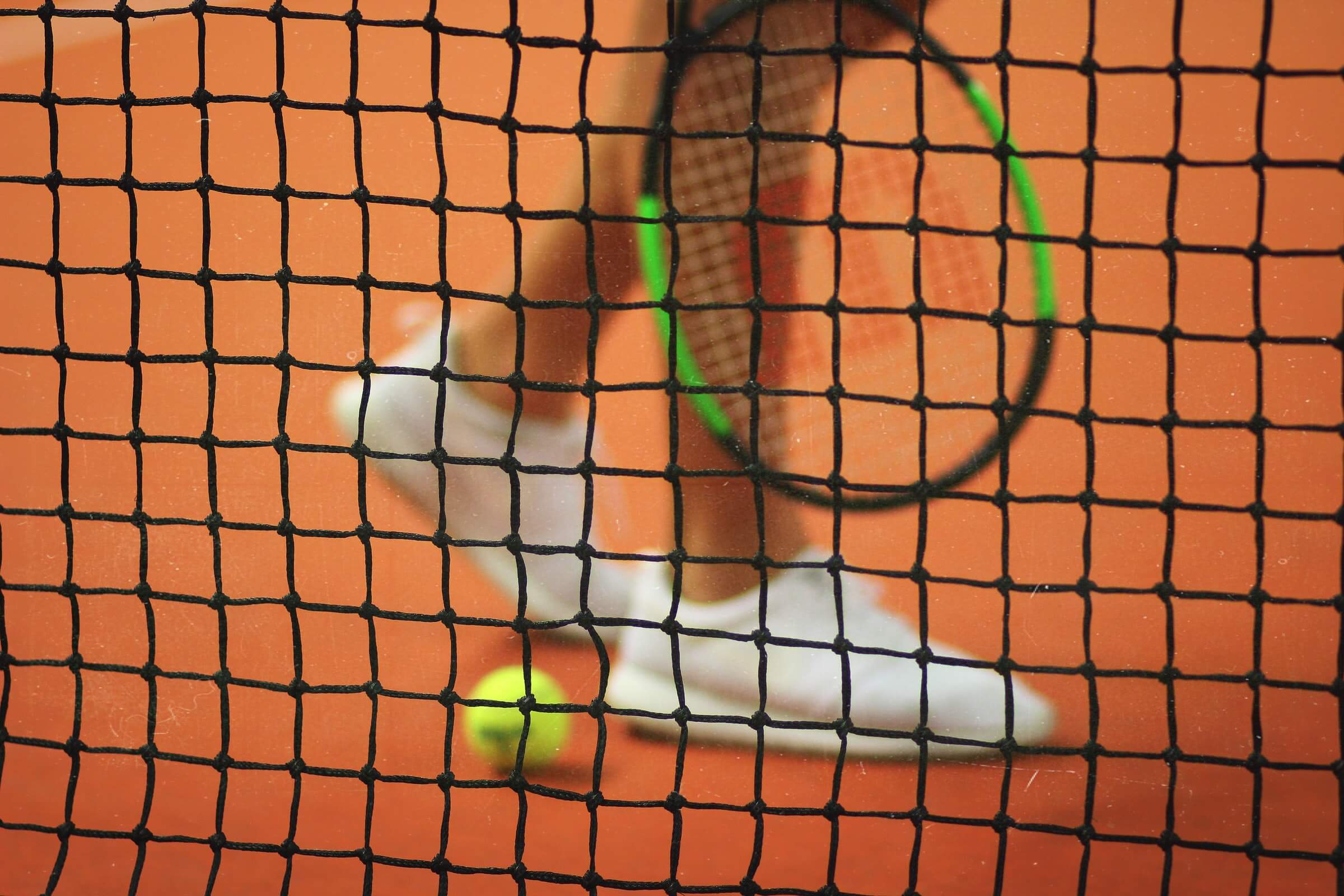 Tennis players legs, ball and racquet seen through tennis netting